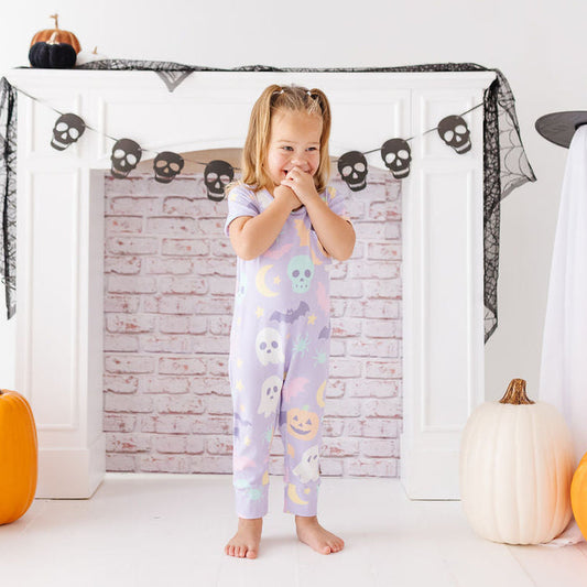 A young girl stands barefoot in front of a white fireplace mantel decorated for Halloween, wearing a light lavender one-piece pajama with colorful ghost and pumpkin prints. White ghost costumes, black netting, skull garlands, and orange pumpkins surround her.
