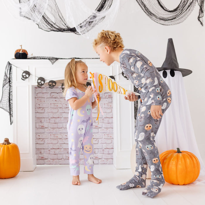 Two children wearing matching Halloween-themed pajamas pose in front of a decorated fireplace. The child on the right wears a gray pajama set with a ghost and owl pattern, and the child on the left wears a lavender pajama set with a moon and owl pattern.