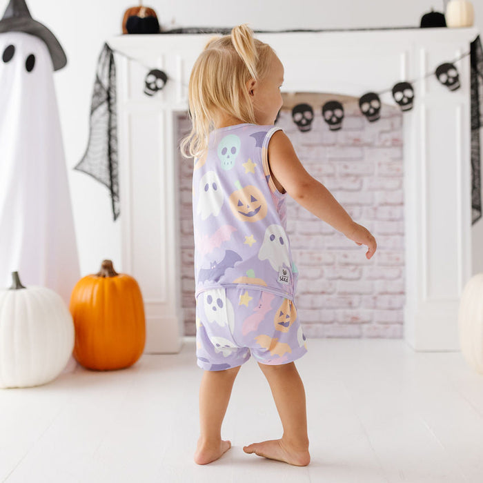 A toddler stands barefoot in front of a fireplace wearing a sleeveless pajama top and shorts set with a pastel ghost and pumpkin print. The room is decorated for Halloween with a ghost figure, pumpkins, and black spider banners.