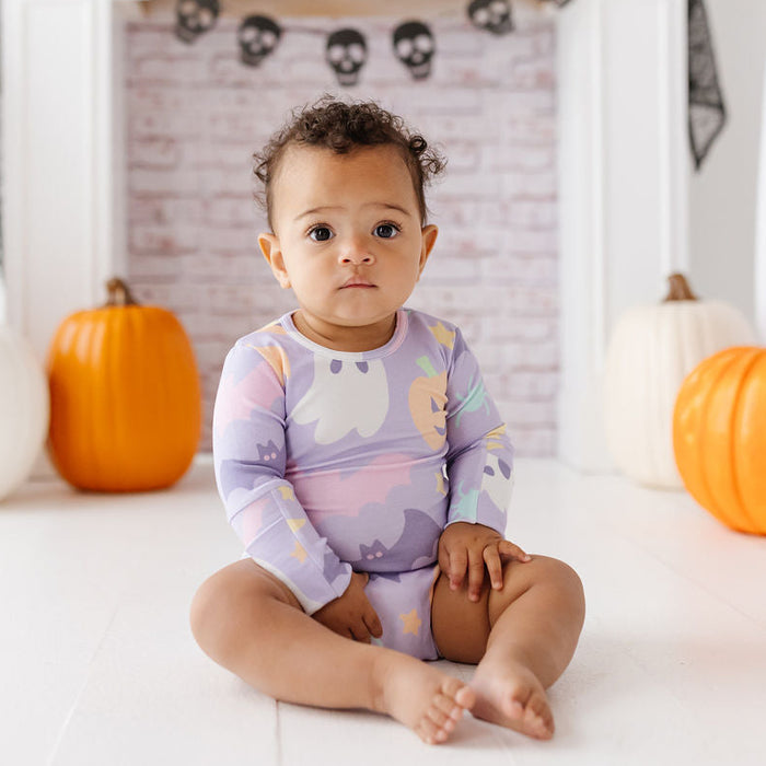 A baby sitting on a white floor in front of a fireplace, wearing a long-sleeve pajama top with a pastel ghost and moon pattern. The room is decorated for Halloween with pumpkins, skull garlands, and black spiderwebs.