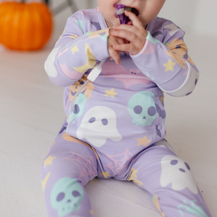 A baby wearing a light purple long-sleeve footed sleeper with a Halloween print of ghosts, skulls, and stars, sitting on a white floor with a blurred orange pumpkin in the background.