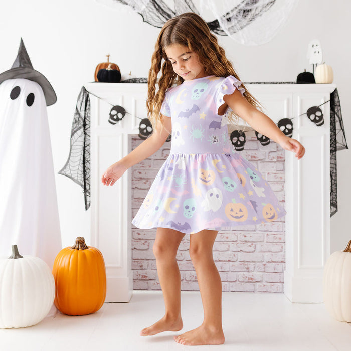 A young girl wearing a lavender short-sleeve Halloween dress with a pumpkin and ghost print, standing barefoot in front of a white fireplace mantel decorated for Halloween.