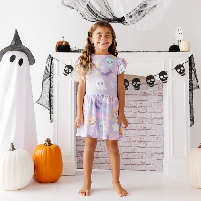 A young girl stands barefoot in front of a white fireplace mantel decorated for Halloween, wearing a short flutter-sleeve dress with a pastel multicolor print of ghosts, pumpkins, and bats. Large paper ghost props, black webbing, and pumpkins are arranged around her in a bright, festive room.