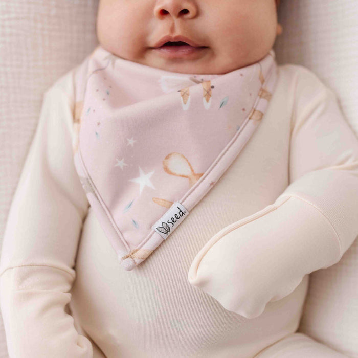 Baby wearing a pink bib with a brand label, sitting on a soft surface.