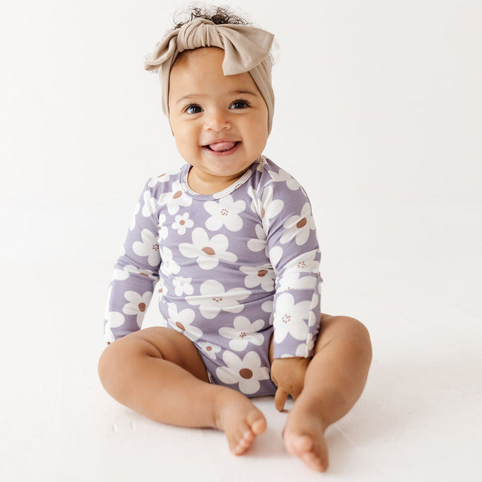 A smiling baby sitting on a white studio floor, wearing a long-sleeve floral romper in lavender and white with a beige bow headband.