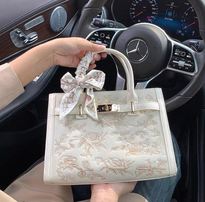 Cream embroidered Kelly-style handbag with gold hardware and a floral bow accent on handle.