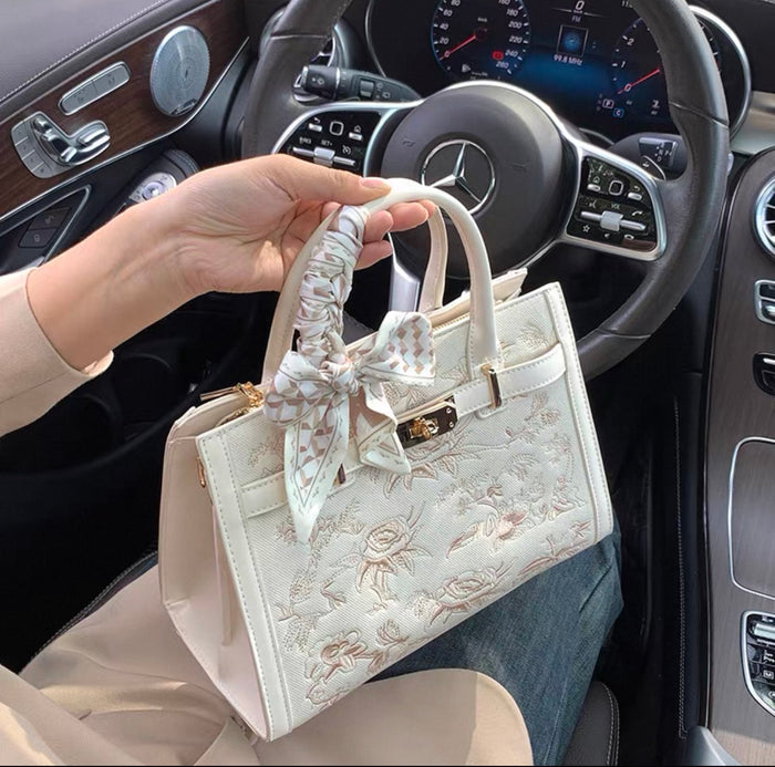 White Kelly-style handbag with floral embroidery and silk scarf tied on handle.
