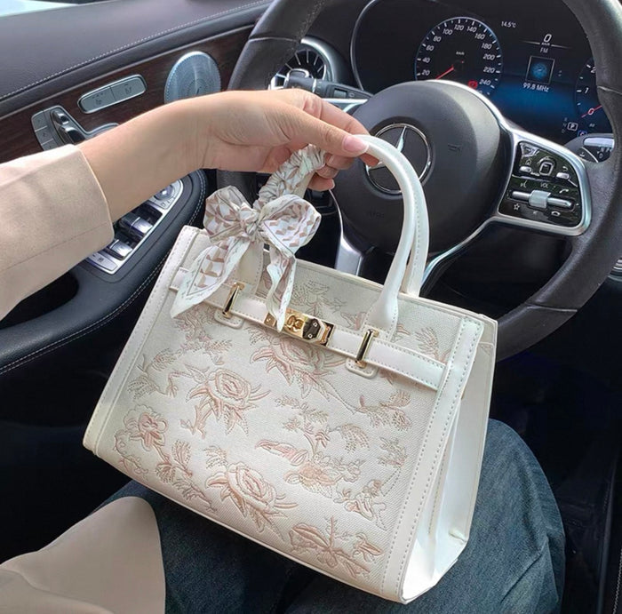 White Kelly-style handbag with floral embroidery, gold hardware, and structured top handle.