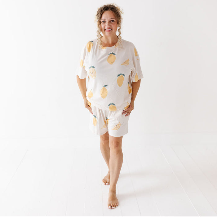 A woman modeling a matching short-sleeve pajama set with a lemon print, standing barefoot against a bright white studio background.