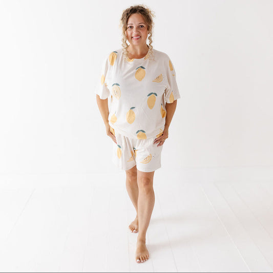 A woman modeling a matching short-sleeve pajama set with a lemon print, standing barefoot against a bright white studio background.