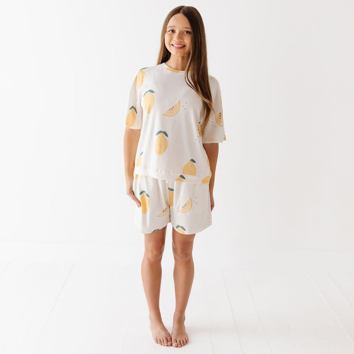 A woman modeling a matching short-sleeve pajama set with a lemon print, consisting of a cream-colored top and shorts, against a clean white studio background.