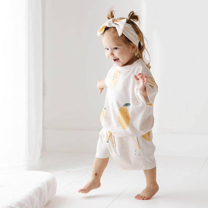 A toddler walking barefoot in a bright, minimalist room, wearing a matching white two-piece outfit with yellow lemon prints and a white bow headband.
