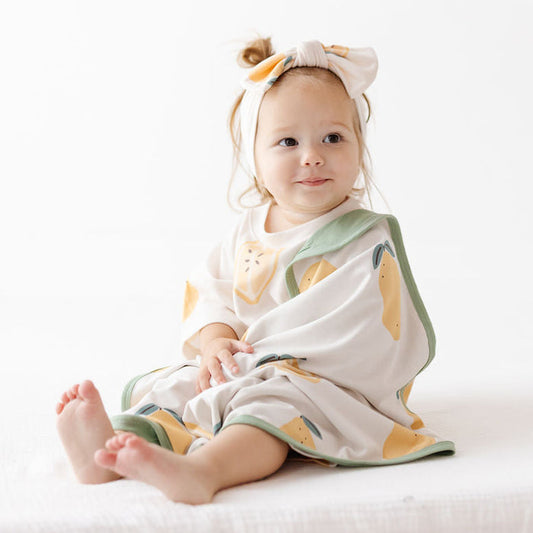 A baby girl sitting on a white fabric surface, wrapped in a white muslin blanket with a yellow and green citrus print, wearing a matching white bow headband.