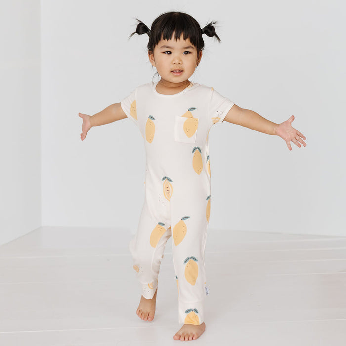 A toddler girl with black pigtails stands barefoot in a white one-piece jumpsuit with a yellow and green lemon print, posing with her arms outstretched in a bright white studio setting.