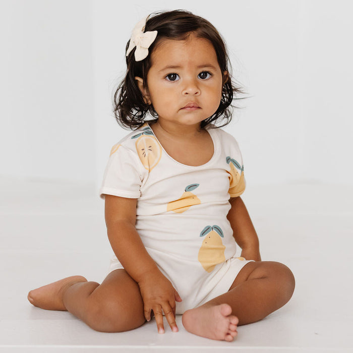 A toddler girl sitting cross-legged on a white floor against a white wall, wearing a white short-sleeve romper with a yellow and green lemon print and a white bow in her hair.