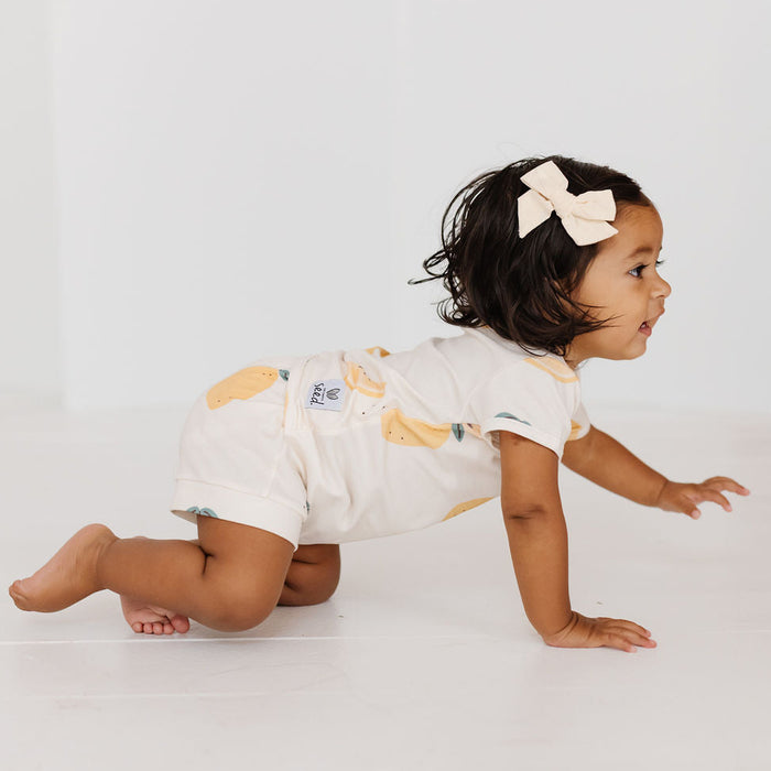 A toddler girl crawling on a white floor wearing a cream short-sleeve romper with a yellow banana print and a matching cream bow headband.