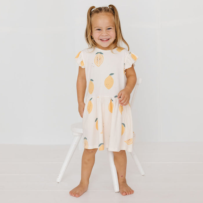 A young girl with blonde pigtails stands barefoot on a white floor in front of a white wall, wearing a cream-colored dress with a yellow lemon print and holding onto a white chair.