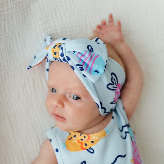 A baby lying on a textured white blanket, wearing a matching light blue outfit set with a bow headband and bib. The set features a playful print of colorful fish, butterflies, and abstract shapes in navy, pink, yellow, and orange.