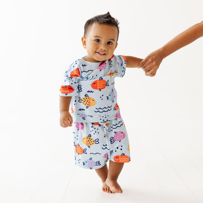 A toddler wearing a light blue two-piece pajama set with a colorful fish and octopus print, walking while holding an adult's hand against a white studio background.
