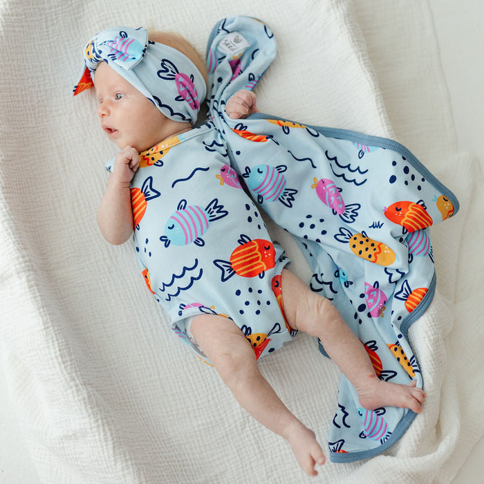A baby lying on a white textured blanket, wearing a light blue fish-print bodysuit, matching hat, and a matching fish-patterned swaddle blanket.