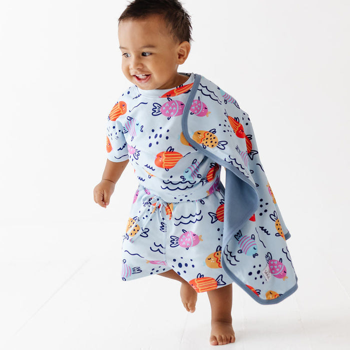 A smiling toddler walking barefoot on a white studio floor while wearing a light blue wrap dress with an orange, pink, and navy ocean-fish print. A matching light blue towel is draped over one shoulder.