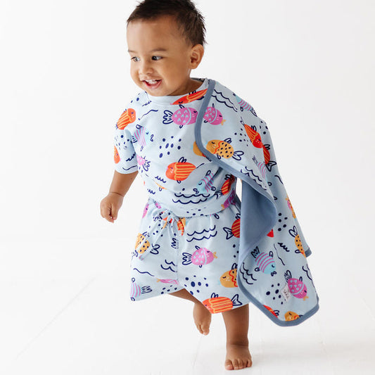 A smiling toddler walking barefoot on a white studio floor while wearing a light blue wrap dress with an orange, pink, and navy ocean-fish print. A matching light blue towel is draped over one shoulder.