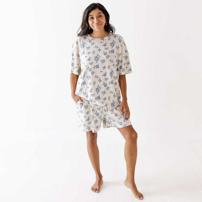 A woman modeling a matching short-sleeve pajama set with a blue floral print on a cream background, standing against a clean white studio backdrop.
