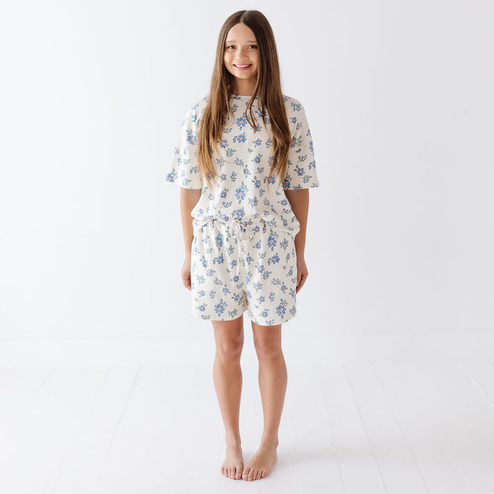 A young girl with long brown hair stands barefoot against a white studio background, wearing a short-sleeve cream-colored romper with a blue floral pattern.