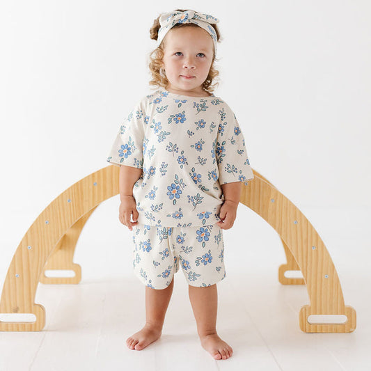 A toddler stands barefoot in front of a wooden arch play structure, wearing a matching cream two-piece outfit with a blue floral print and a coordinating floral headband.