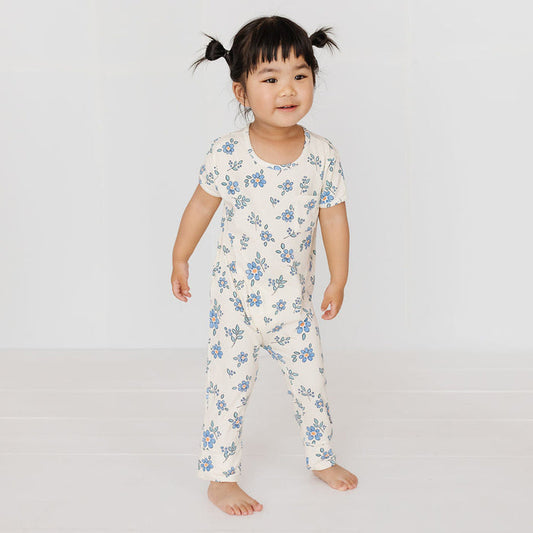 A toddler girl with dark hair in two pigtails stands barefoot in a white one-piece jumpsuit with a blue floral pattern against a clean white studio background.
