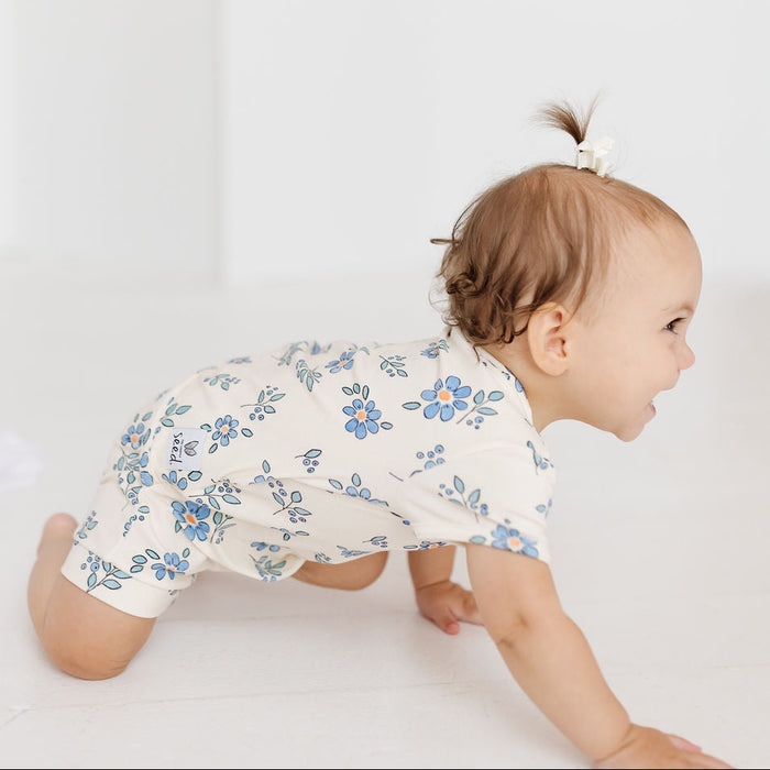 A baby crawling on a white floor wearing a cream-colored short-sleeve romper with a blue floral pattern. The baby has a small hair bow in their hair and is looking to the right.