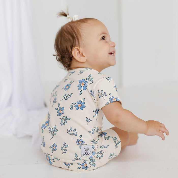 A baby sitting on a white floor, wearing a cream-colored short-sleeve romper with a blue floral pattern. The baby is shown from a side-back angle, looking to the side with a smile.