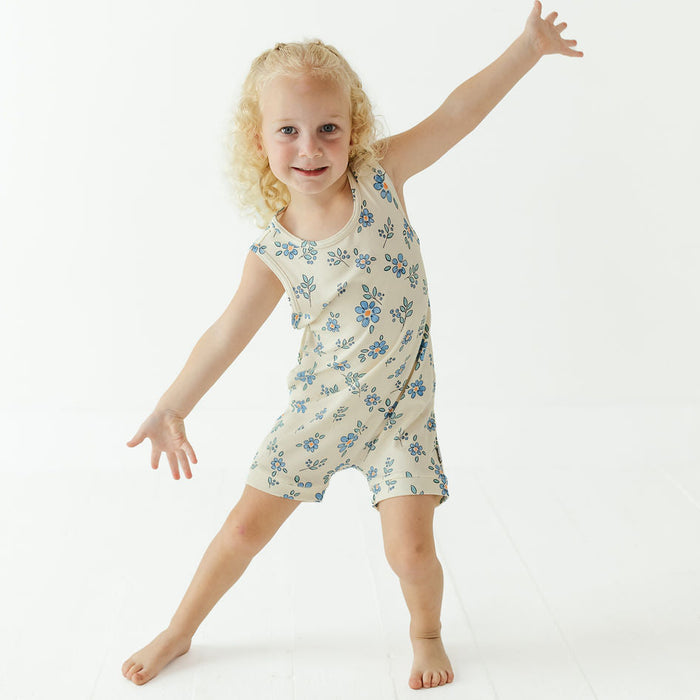 A young blonde girl posing in a sleeveless cream-colored romper with a blue floral pattern against a bright white studio background.