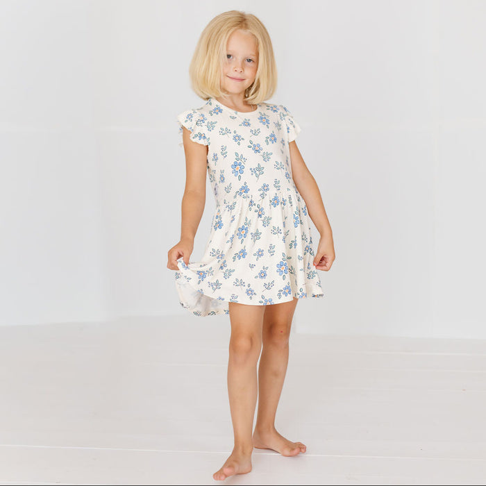 A young blonde girl stands barefoot against a plain white studio background, wearing a short-sleeve white dress with a light blue floral print.