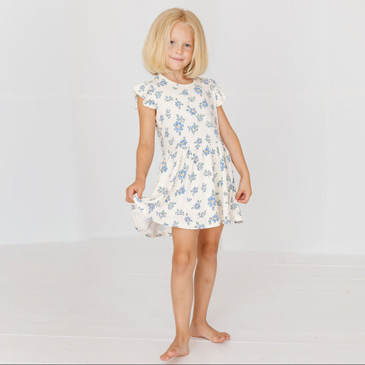 A young blonde girl stands barefoot against a plain white studio background, wearing a short-sleeve white dress with a light blue floral print.