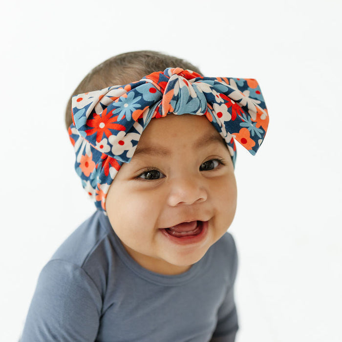 A baby wearing a blue long-sleeve bodysuit and a colorful floral bow headband, smiling against a plain white background.