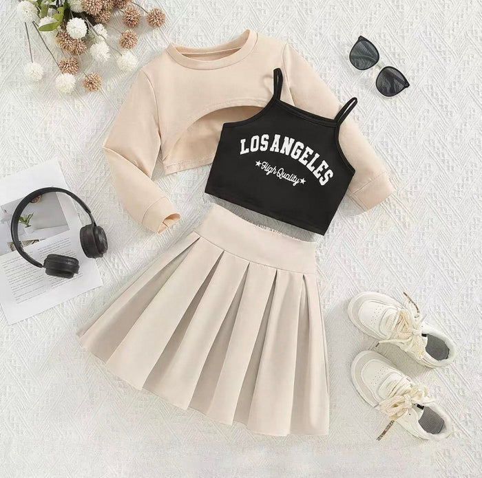 Beige long-sleeve top, black 'Los Angeles' crop top, beige pleated skirt, white sneakers, sunglasses, and headphones on a textured white surface.