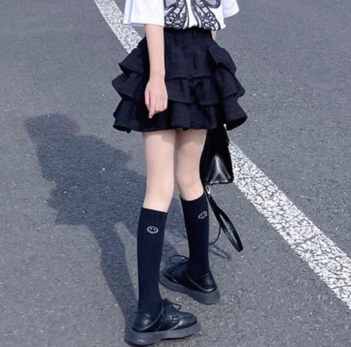 Person wearing a black layered skirt with knee-high black socks and black shoes on a gray pavement.