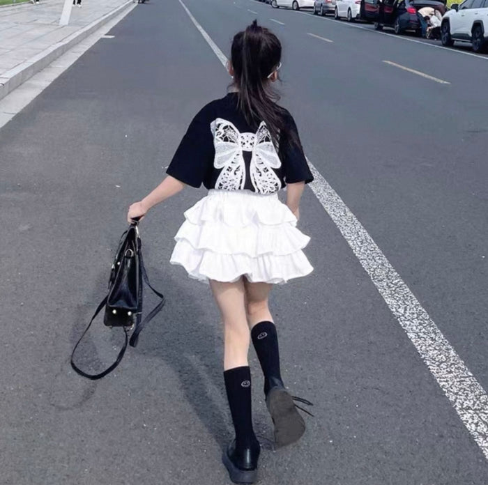 Person walking on a road wearing a black t-shirt with a butterfly design, white skirt, and black knee-high socks.