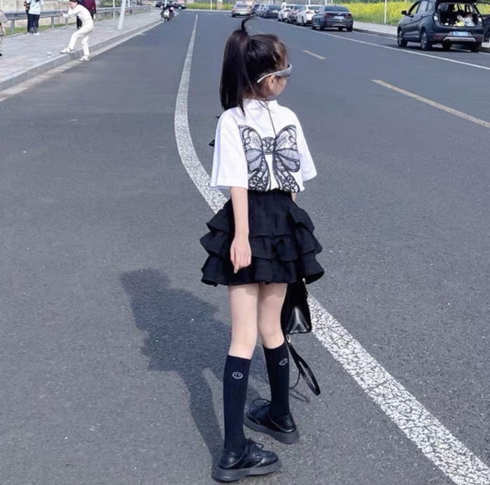 Person wearing a white top with butterfly design and black ruffled skirt on a road.