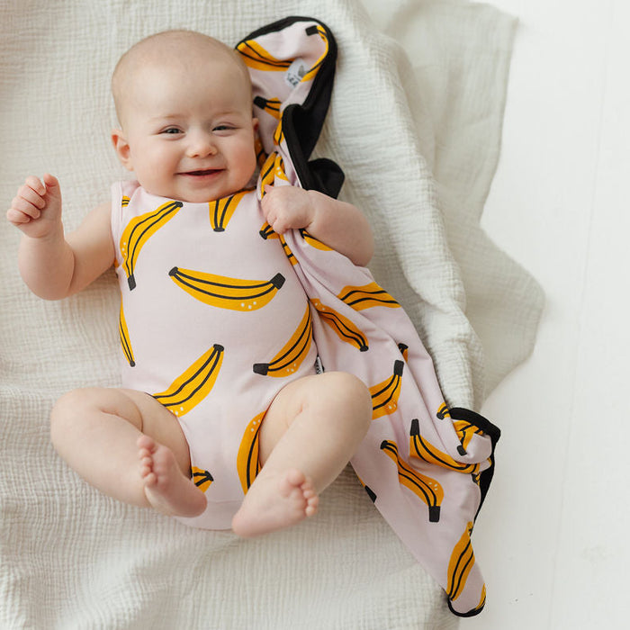A baby lying on a white muslin blanket, wearing a light pink sleeveless bodysuit and holding a matching pink blanket with a yellow banana print.