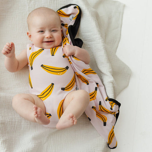 A baby lying on a white muslin blanket, wearing a light pink sleeveless bodysuit and holding a matching pink blanket with a yellow banana print.