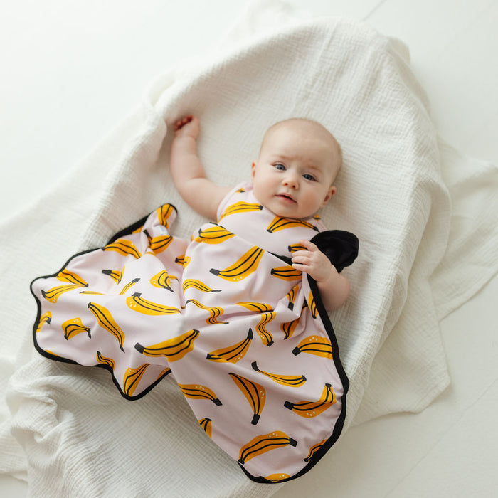 A baby lying on a white textured blanket, wrapped in a light pink muslin swaddle blanket with a yellow banana print and black trim.
