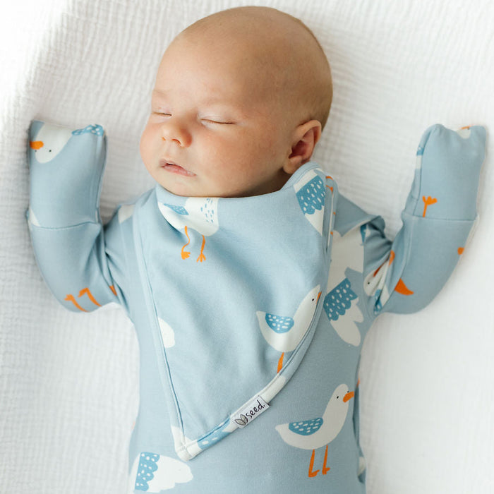A sleeping newborn baby wearing a light blue long-sleeve sleep suit with a bib and matching foot mittens, printed with a white and blue seagull pattern, lying on a white textured blanket.