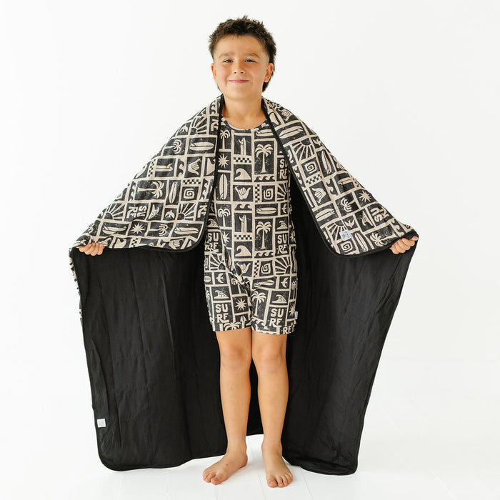 A young boy stands barefoot against a white studio background, holding open a large black towel or blanket with a white geometric pattern draped over his body like a cape. He is wearing a matching black and white patterned towel or blanket draped over his body.