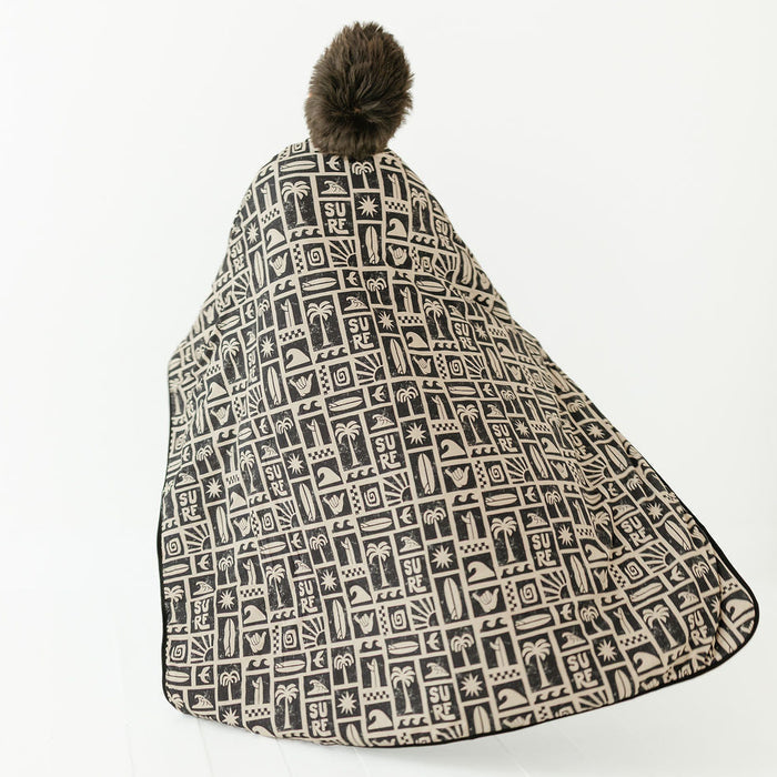 blanket with all-over black and beige graphic pattern, featuring a dark brown faux-fur pom-pom on the hood, shown against a plain white background.