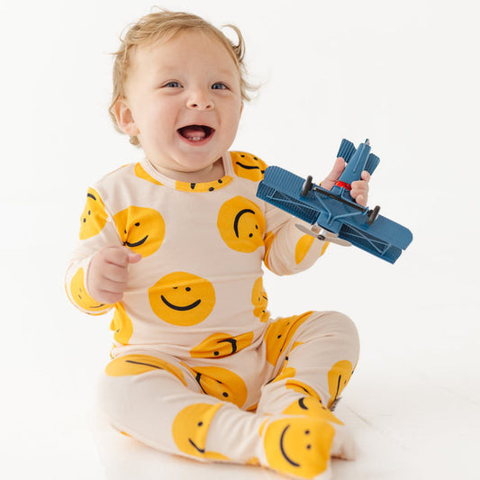 Child wearing a smiley face onesie holding a blue toy airplane on a white background