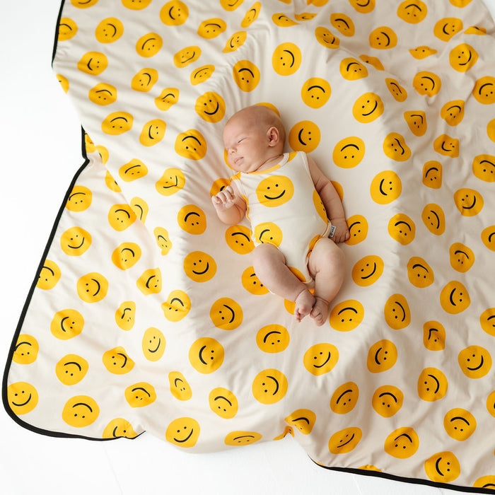 A sleeping newborn baby wearing a cream bodysuit with a yellow smiley face pattern, lying on a matching cream baby blanket with a black border and yellow smiley face print.