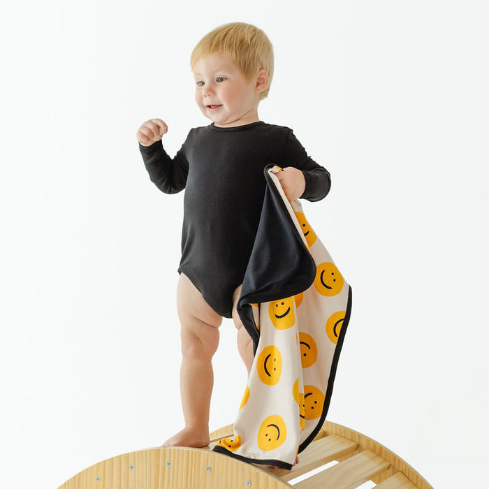 A toddler stands on a wooden arch in a studio setting, wearing a black long-sleeve bodysuit and holding a white muslin cloth with a yellow smiley face pattern.