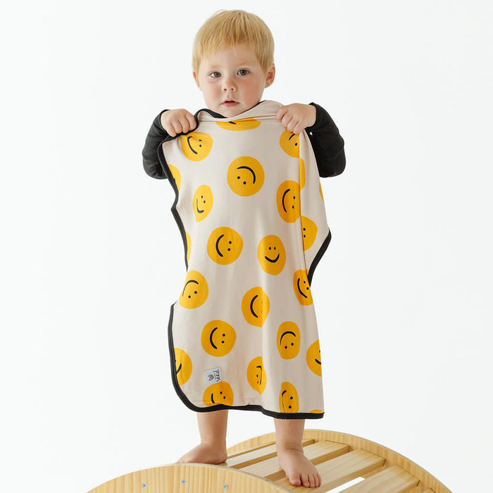 A baby wearing a black long-sleeve shirt stands on a wooden arch and holds up a cream-colored blanket with a yellow smiley face pattern.